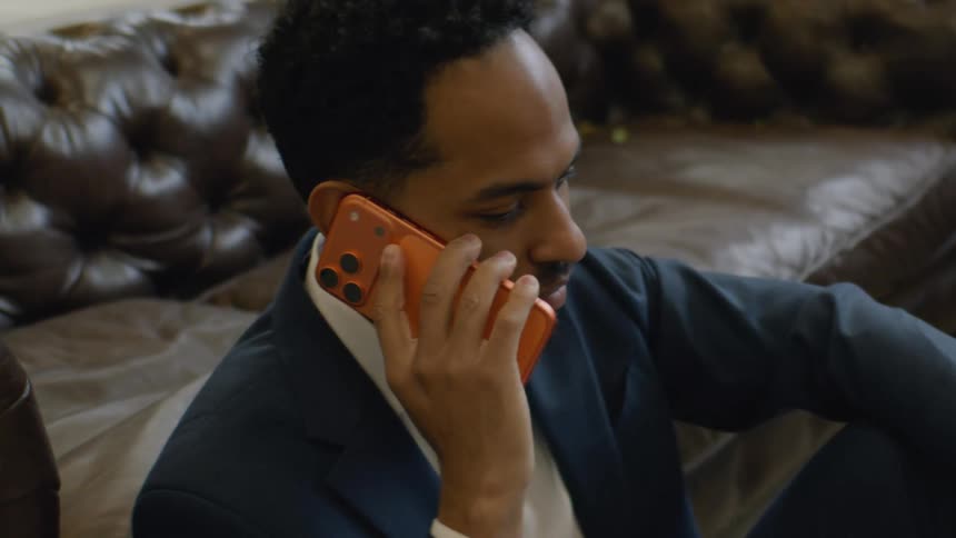 A young man sits on the floor in his living room at midday leaning back against a sofa hes wearing a blue blazer suit and holding an iphone 17 to his ear as he makes a phone call