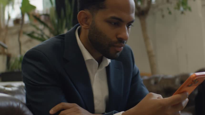 Man seated on a sofa using a smartphone indoors with plants in the background