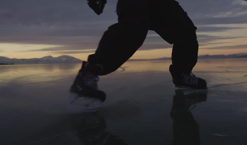 A person wearing skates and holding a hockey stick skates across a frozen lake toward the camera during a sunset with mountains in the background
