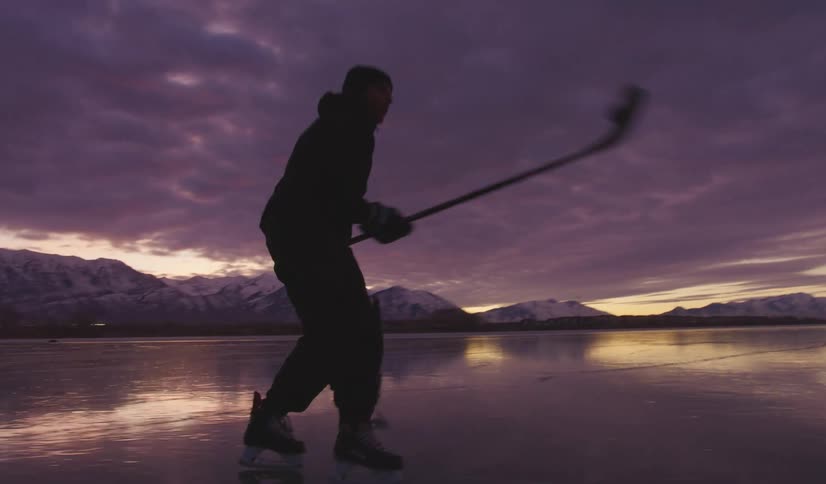 Person skating and handling a hockey puck on a frozen lake at dusk with mountains in the background