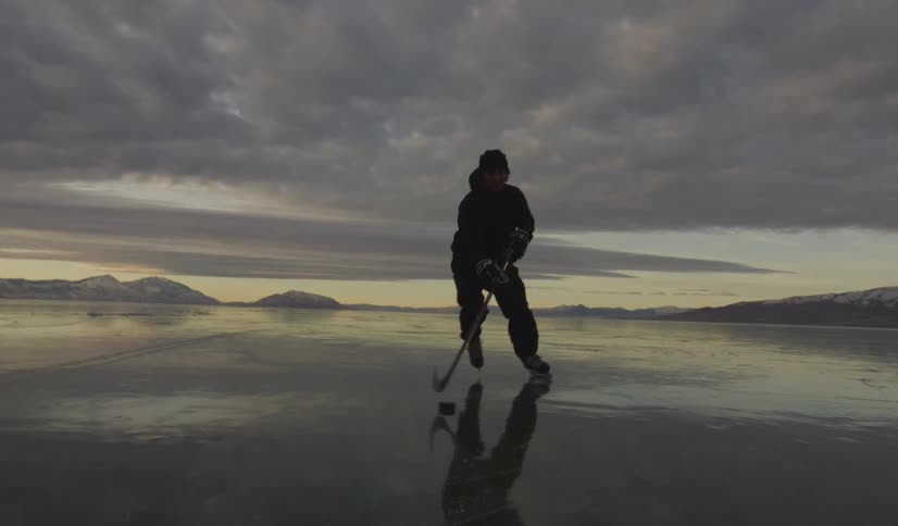 A man skates across a vast frozen lake while handling a hockey stick under a cloudy twilight sky