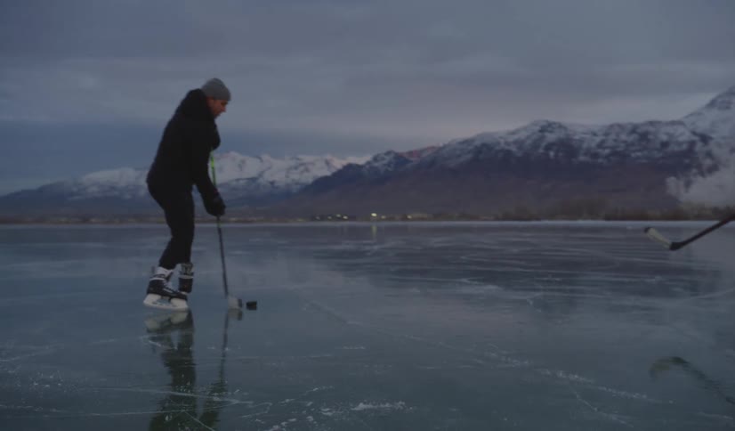 Two men in black athletic wear play ice hockey on a vast frozen lake during twilight maneuvering a puck with sticks against a backdrop of massive snowcovered mountains and a dim sky