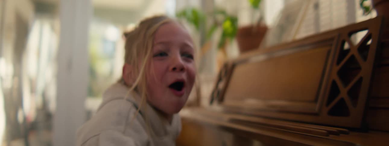 A young girl with blonde hair tied in a bun laughs joyfully while playing a wooden upright piano in a sunlit room