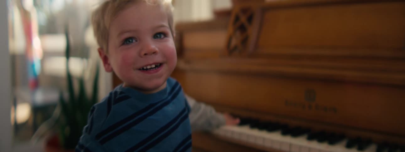 A young boy with blonde hair and blue eyes smiles and laughs while standing in front of a wooden piano