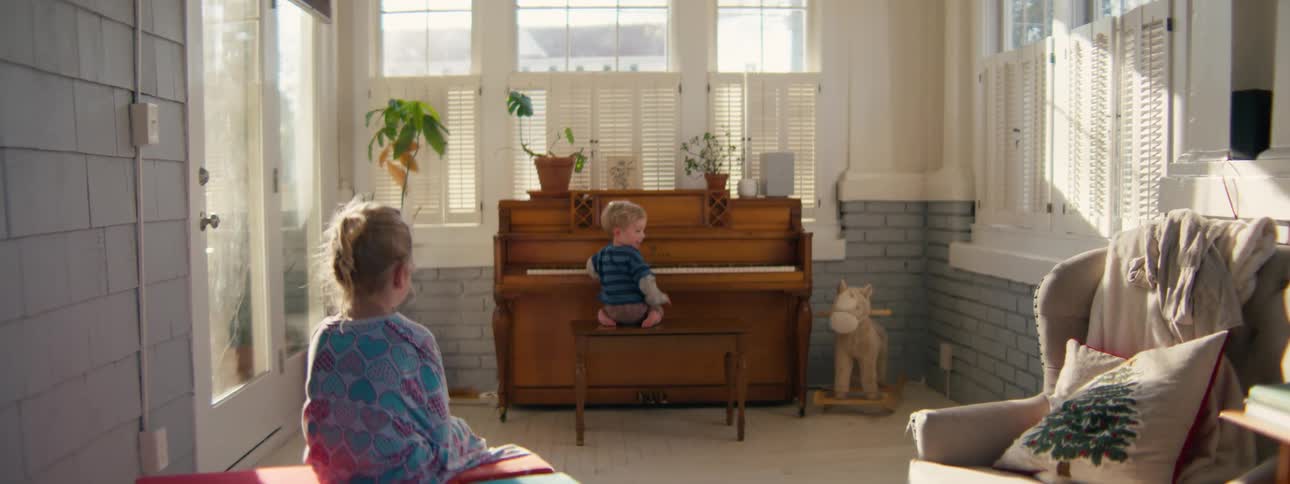 Male toddler sitting and playing the piano then looking back and smiling to a female child sitting on a bed watching him play indoors during day time