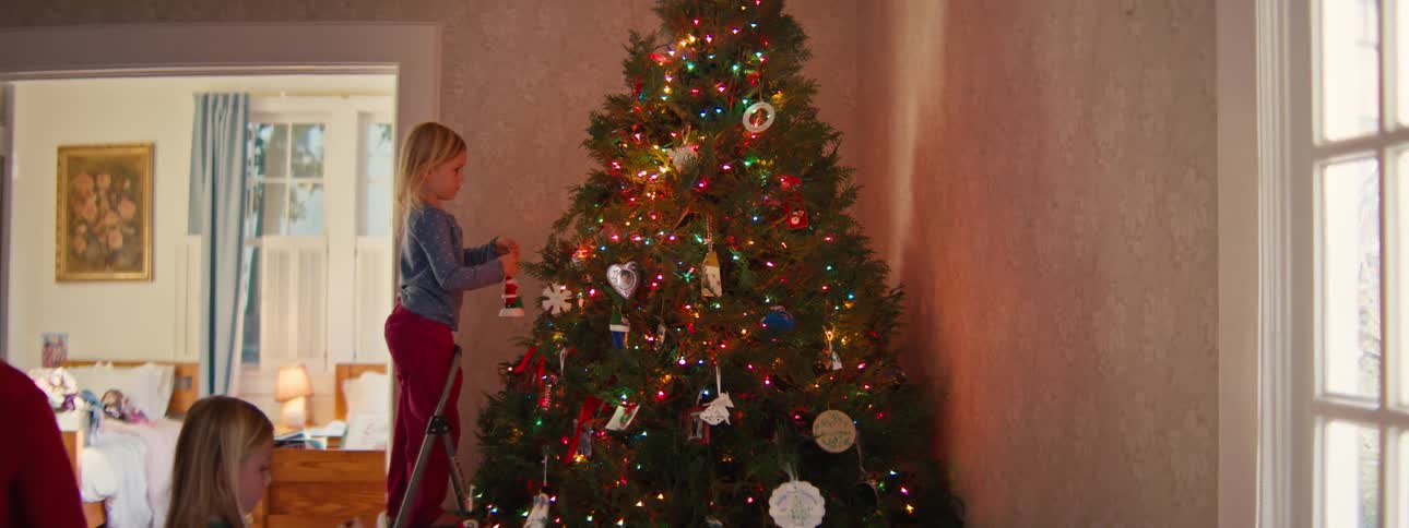 A young girl stands on a small stepladder to hang an ornament on a brightly lit christmas tree while another child sits nearby in a cozy living room