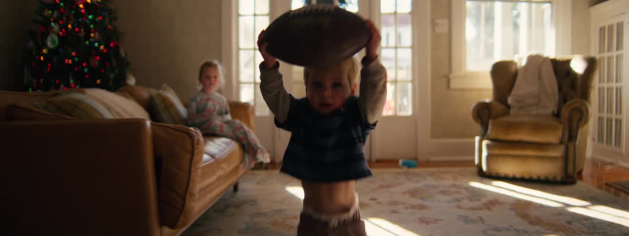 A toddler boy excitedly runs toward the camera through a sunlit living room while holding a large football above his head as a young girl watches from a leather sofa near a decorated christmas tree