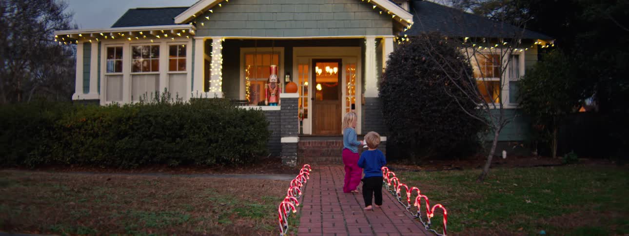 A toddler and a young girl walk along a decorated walkway at sunset heading toward a house with christmas lights and ornaments glowing softly in the warm evening light
