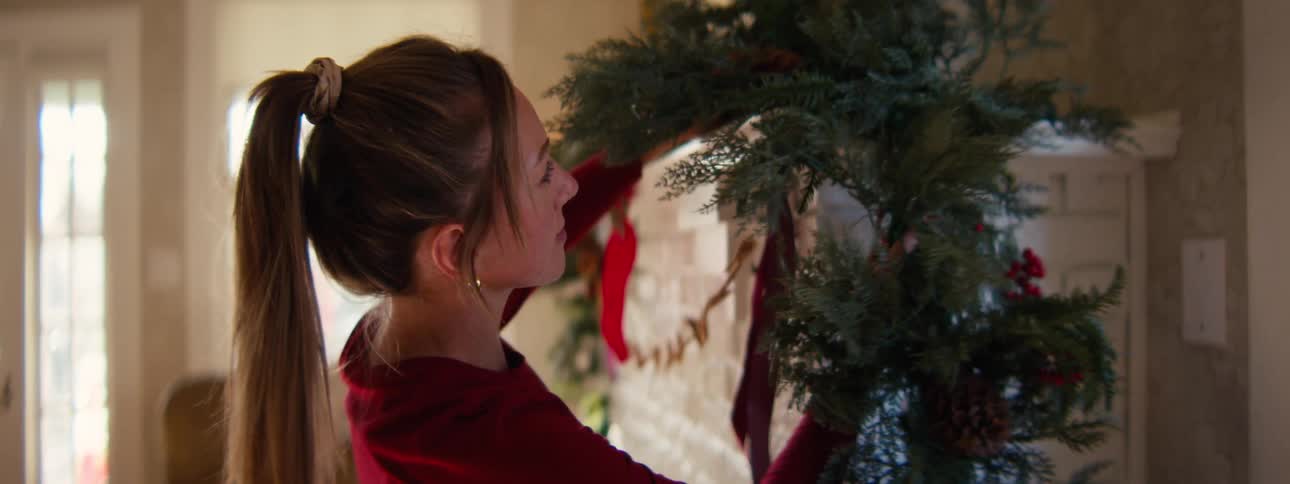 A woman in a red shirt decorates a white brick fireplace with a green garland