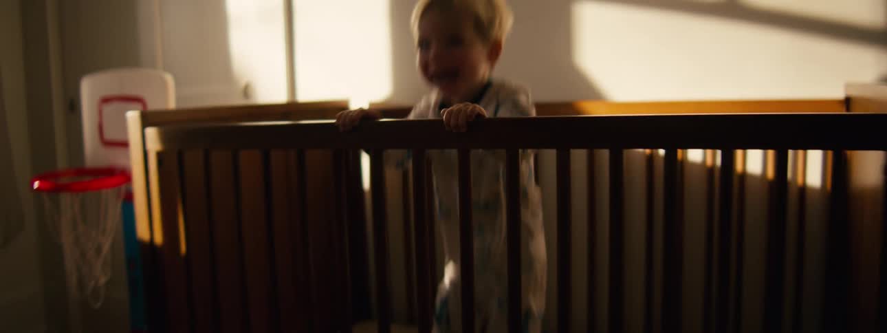 A toddler in patterned pajamas stands and jumps excitedly inside a wooden crib while golden sunlight filters into the room showing a small basketball hoop nearby