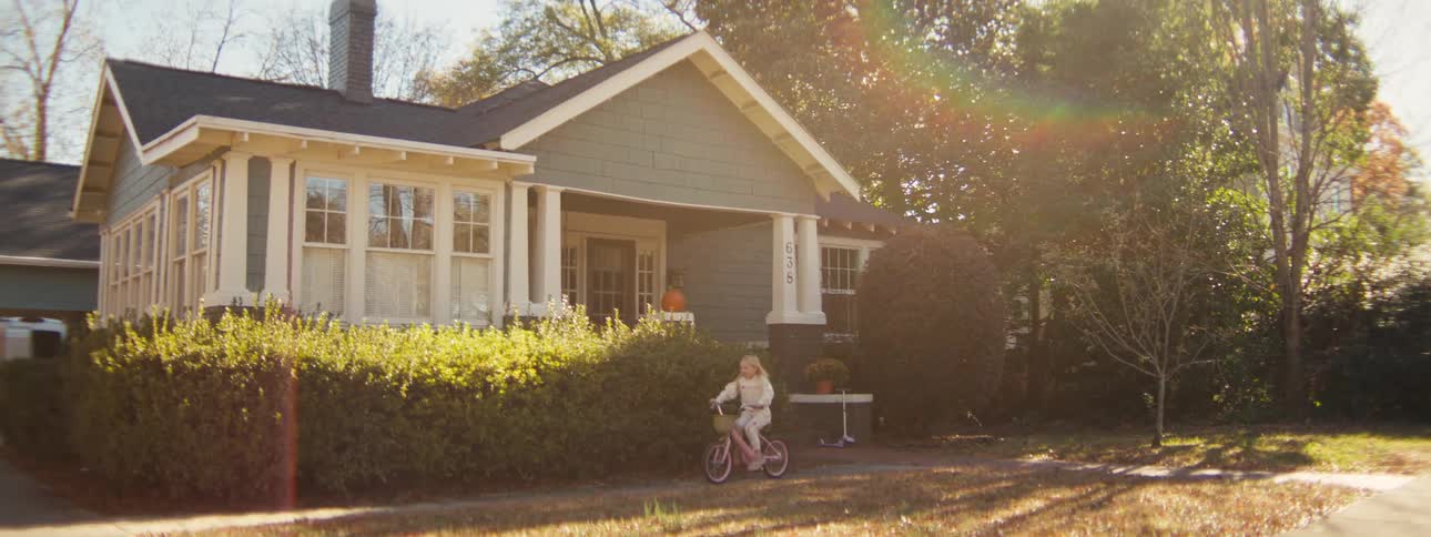 A young girl with blonde hair rides a pink bicycle down a sunlit sidewalk in front of a house