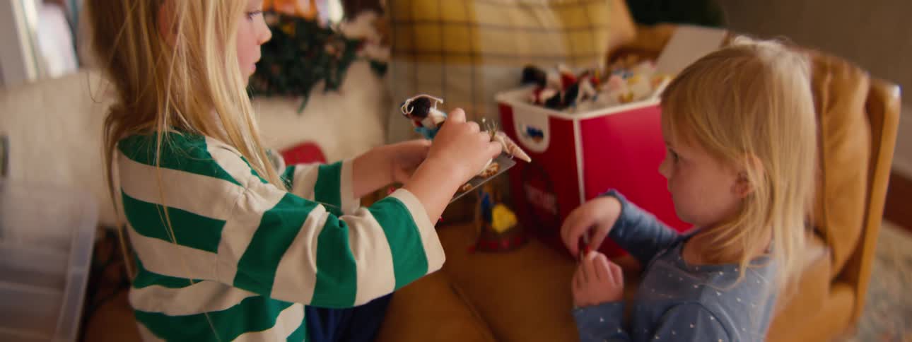 Two young blonde sisters sit together on a tan leather couch focused on playing with small toy figurines and a red storage bin while a blurred christmas tree stands in the background of the living room