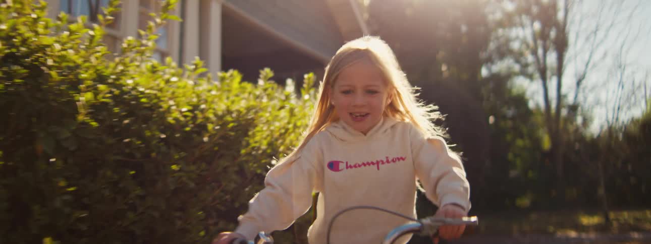 A young girl with long blonde hair rides a bicycle across the front lawn on a sunny day shes wearing a sweatshirt and the bright light makes the grass look warm and green as she moves