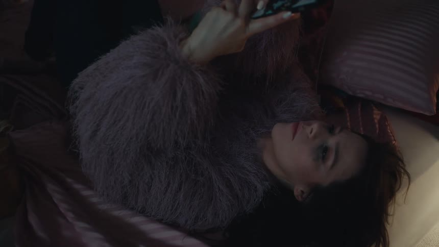 Woman lies on her bed and holds her mobile phone in a dimly lit indoor environment