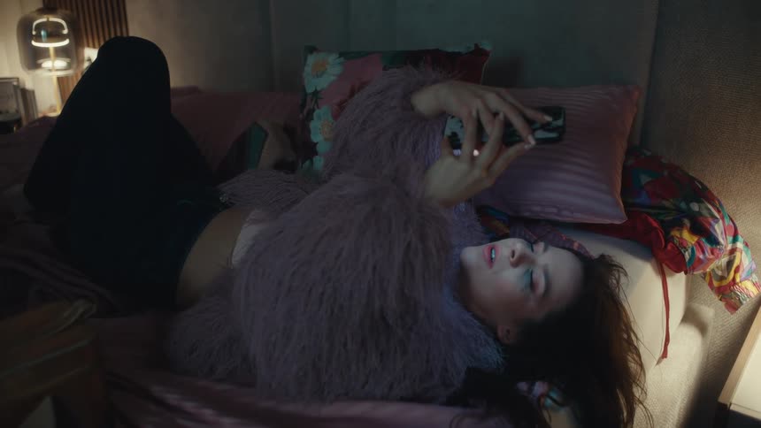 A young woman lies on her bed at night scrolling on her mobile device the room is dim with only the soft glow from the screen lighting her face as she relaxes quietly