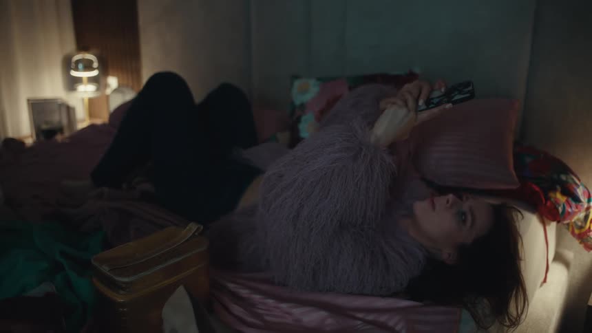 Woman lying on a bed holds a mobile phone in a lowlight indoor setting
