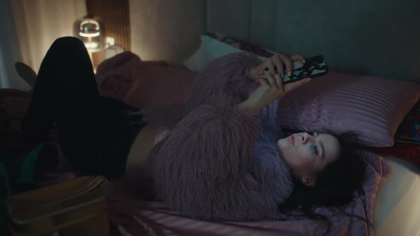 A woman lies on a bed scrolling on a smartphone at night in a softly lit bedroom
