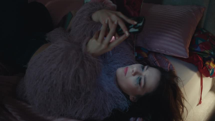 A woman in a purple faux fur jacket walks in a bedroom then lies on a bed and uses a smartphone looking forward with a contemplative mood