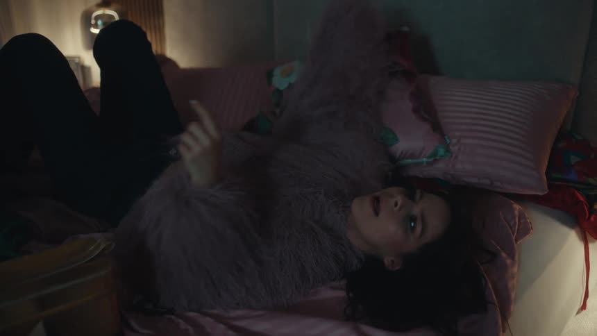 A woman lies on a pink bed using her smartphone before she frustratedly tosses the device aside and covers her face with a blue pillow in a dimly lit bedroom