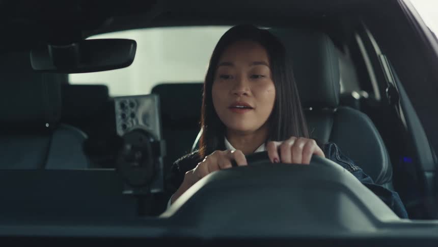 Smiling and talking young adult woman attaching a cell phone to a mount inside a car then reacting in a scared and nervous manner before smiling and talking again