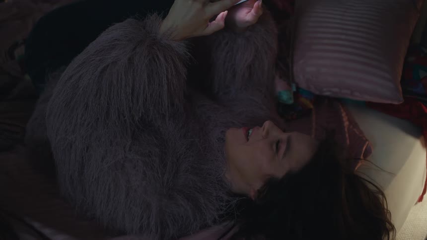 A young woman walks into her room at night then hops onto her bed with a light playful jump she settles onto her back grabs her mobile device and begins scrolling