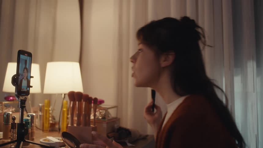 Woman applies makeup while recording a video with a phone on a tripod on a lit vanity