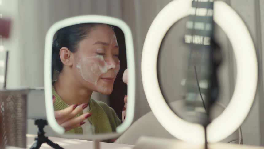 A woman cleans her skin using a brush while recording herself on her smartphone