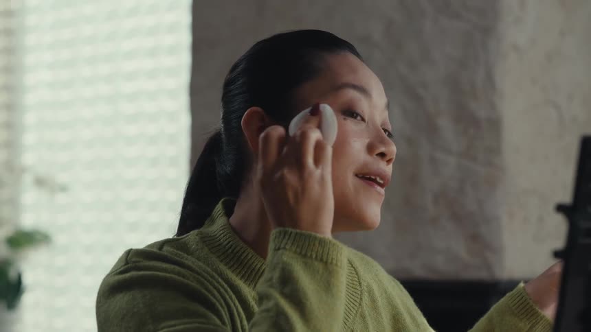 A woman with dark hair wearing a green cardigan uses a cotton pad to apply liquid from a blue bottle to her face while recording herself with a mobile phone