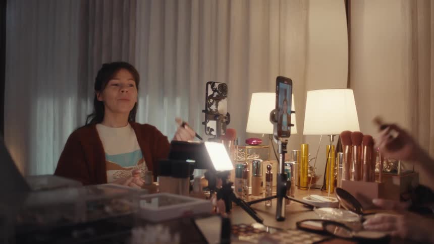 Woman films a makeup tutorial while applying cosmetics at her vanity in a softly lit bedroom