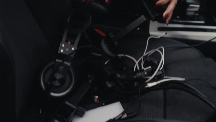 A woman searches through electronic devices and tangled charging cables inside a car