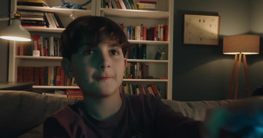 Two young boys are sitting on a couch in a dimly lit room intensely focused on playing a video game the boy in the foreground moves his controller with excitement and determination while bookshelves filled with books and games line the wall behind them