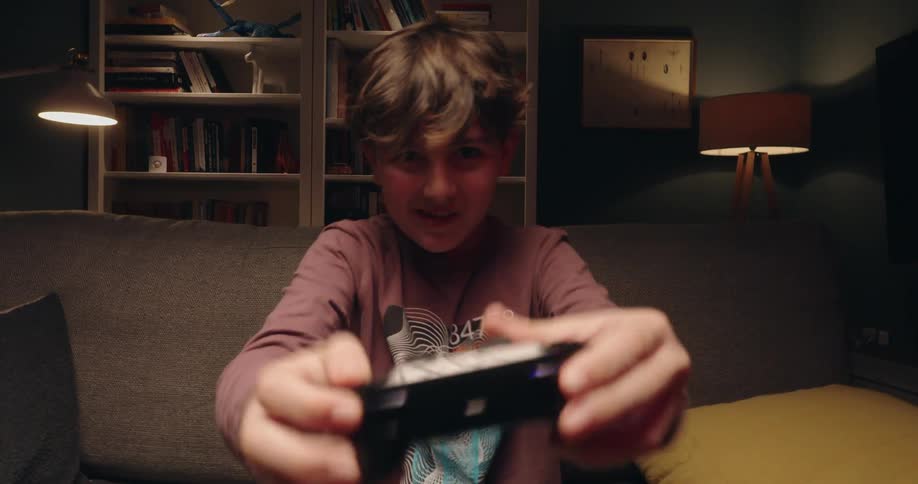 A closeup shot shows a boys hands holding and using a black gaming controller while a persons face is visible in the blurred background