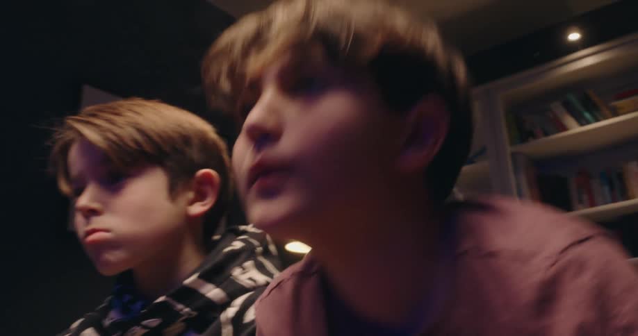 Two boys sit on a couch competitively playing a video game with controllers in a living room