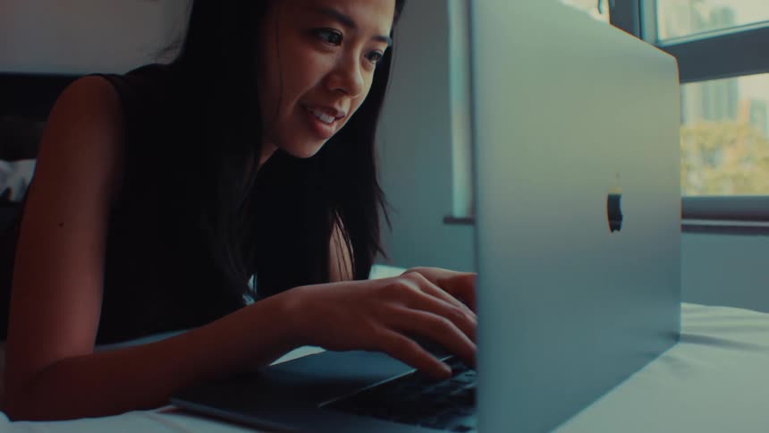A young woman with long dark hair lies on a bed and works on a laptop in a sunlit room
