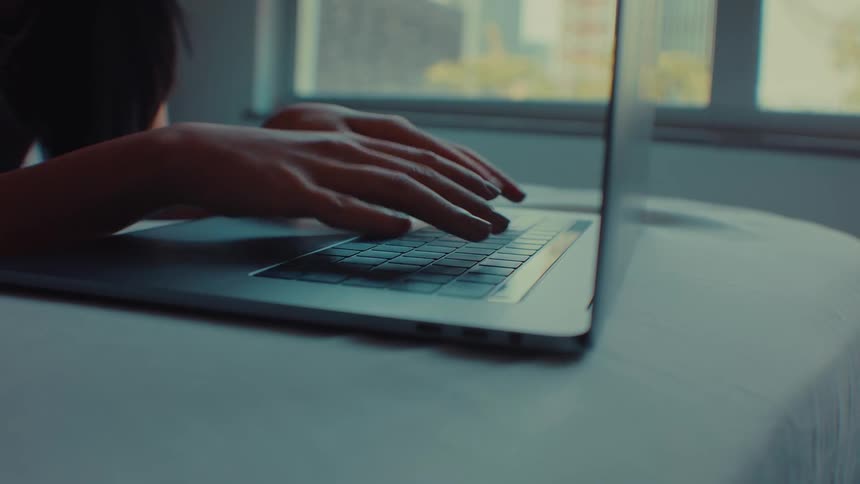 A woman types on the keyboard of her laptop on the edge of her bed