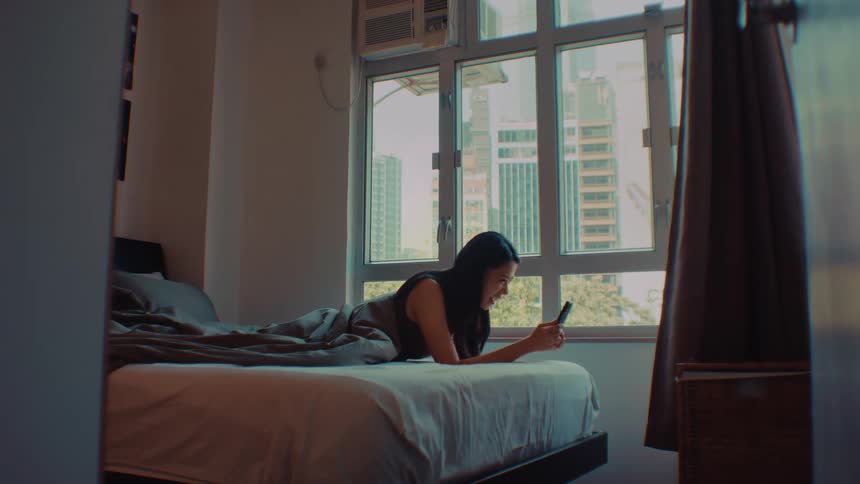 A woman lies on a bed and looks at a smartphone by a bright window in a dimly lit room looking forward with a happy mood