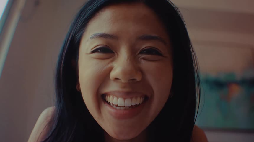 A young woman sits indoors at midday smiling brightly as she waves excitedly during a video conference call sunlight fills the room giving the scene a warm lively feel while she greets the caller with enthusiasm