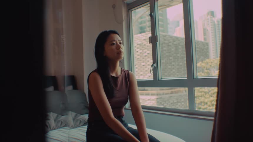 A woman in a sleeveless top sits on a white bed and gazes toward a window in a dimly lit room looking forward with a sad pensive mood