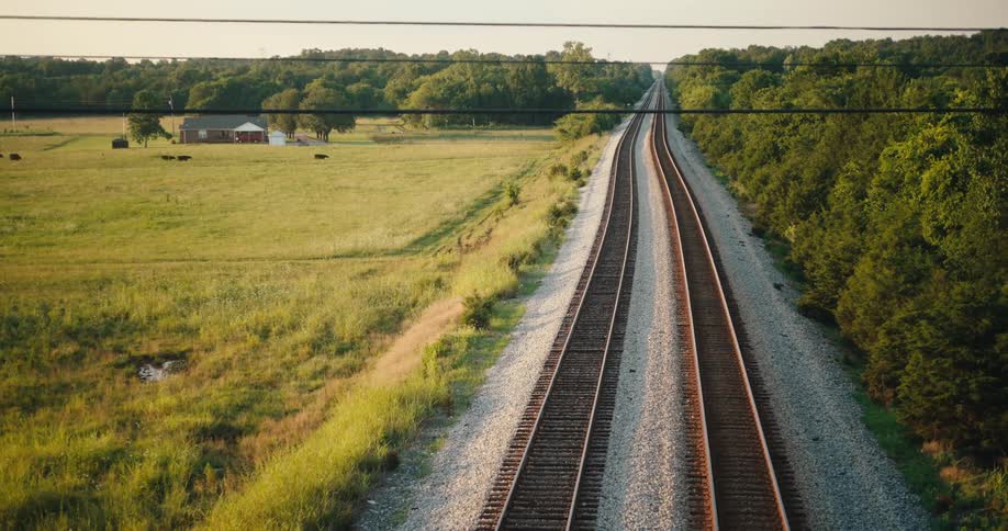 A static highangle shot of a doubletrack railway stretching into the distance through a rural landscape with green fields a house and trees under a clear sky