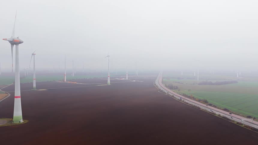 An aerial high angle shot shows a large white wind turbine with red tipped blades spinning slowly over a brown field next to a busy highway