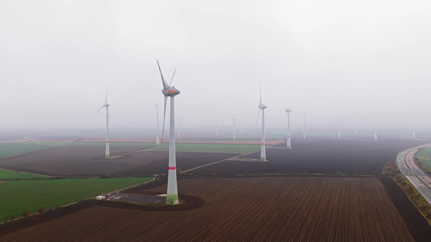 A vast wind farm stretches across flat agricultural fields on an overcast day its tall turbines spinning in unison as cars travel on an adjacent highway