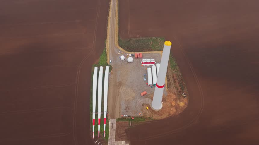 A wind turbine under construction in a plowed field with its blades lying on the ground