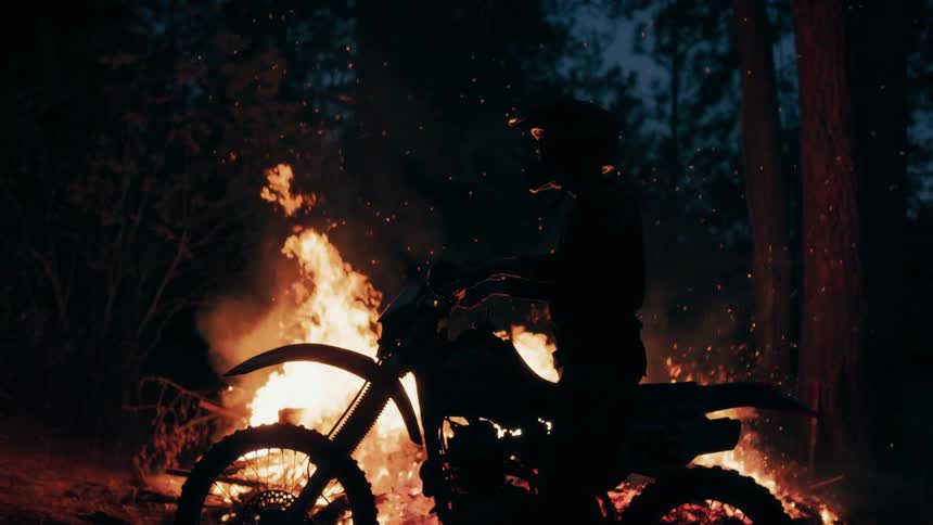 A silhouette of a person in a helmet sits on a dirt bike in front of a massive bonfire at nightglowing embers float through the air as the intense orange flames flicker against the dark forest background
