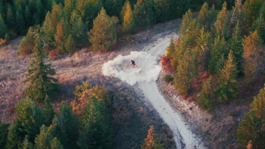 An aerial shot shows a person on a dirt bike performing donuts on a dirt path intersection creating a large cloud of dust