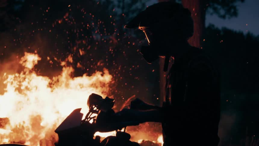 Motorcyclist watches intense fire beside bike in dark area with trees in background