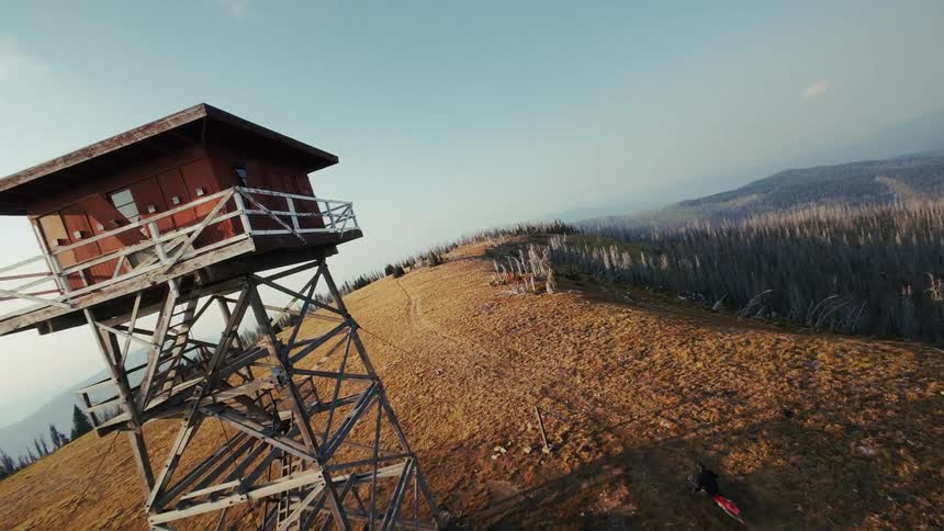 Motorcyclist rides along a path on top of a hill overlooking a conifer forest passing a lookout tower