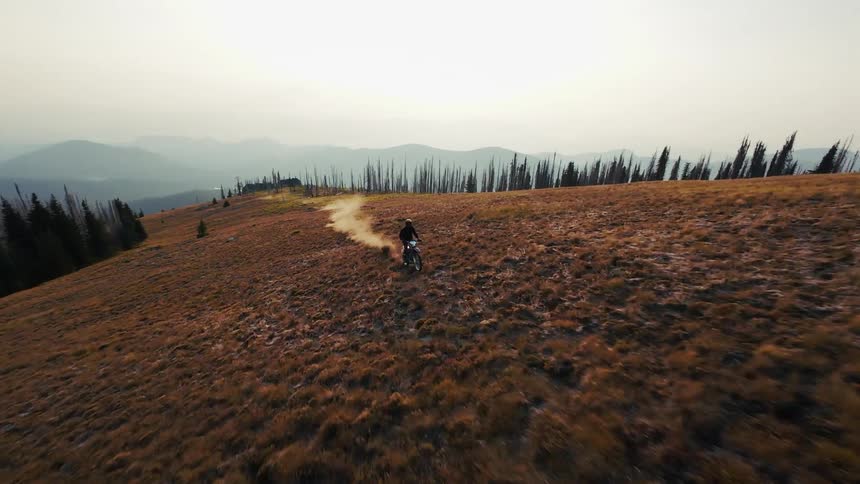 A dirt bike rider accelerates across a wide grassy mountain ridge leaving a thick trail of dust in their wakethe highaltitude landscape features sparse burnt trees and rolling mountain peaks under a hazy sky