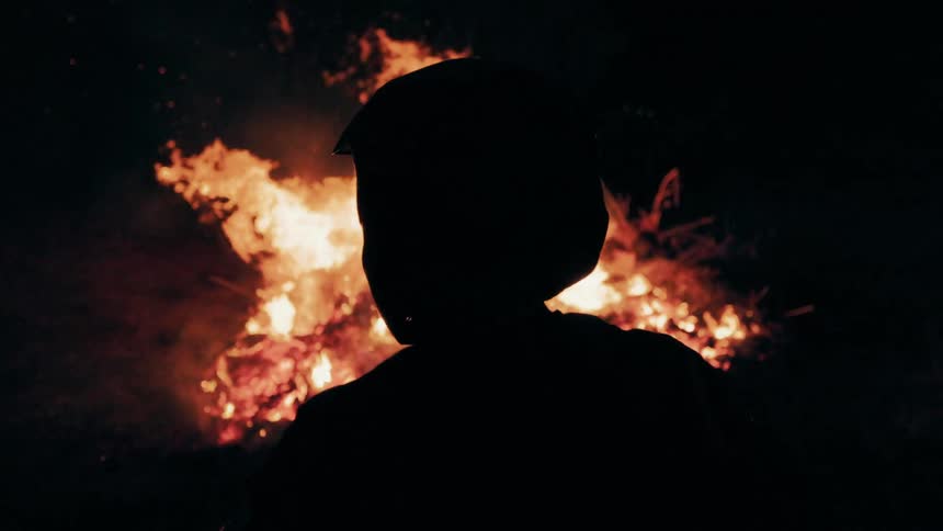 A person wearing a protective helmet and mask is shown in silhouette while facing a large bright fire at nightthe glowing orange flames flicker and dance in the background creating a sharp contrast against the dark foreground