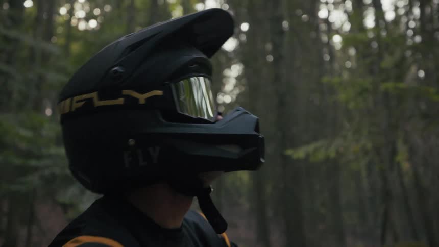 A mountain biker wearing a black and yellow fly racing helmet gazes upward while paused in a dimly lit forestthe riders goggles reflect the surrounding tall trees as they take in the quiet wooded environment