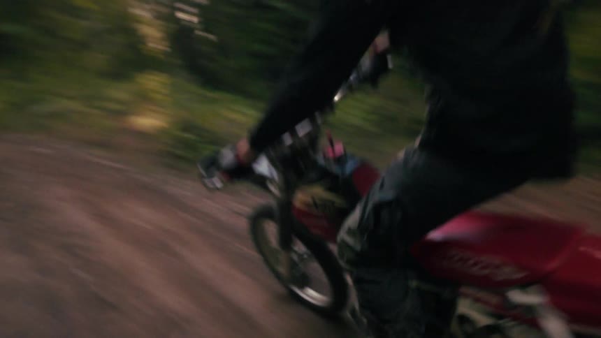 A motorcyclist wearing a black helmet and gear rides a dirt bike quickly down a narrow forested paththe camera follows closely behind as the rider kicks up dust while navigating the winding trail through dense green trees